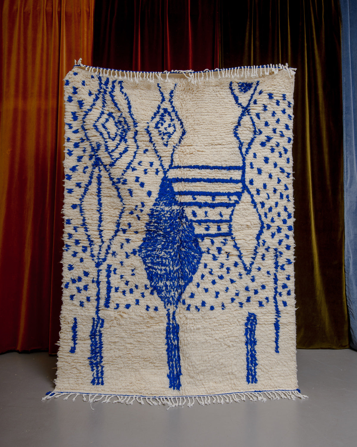 Azilal rug with abstract design in blue with white cotton tassels, being held up in front of a wall of velvet curtains.