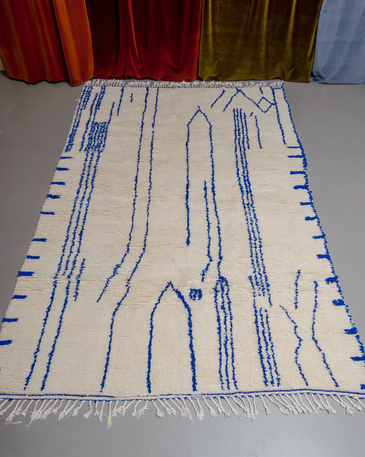 Berberlin Azilal rug in cream wool with blue line pattern and white cotton tassels laid out on grey floor with velvet curtains behind.