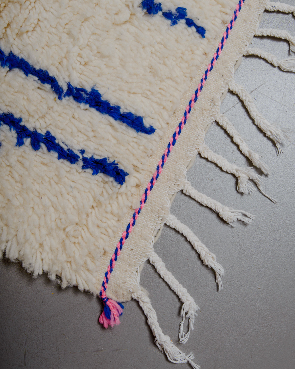 Corner of azilal rug with white cotton tassels and pink accents on grey floor. 