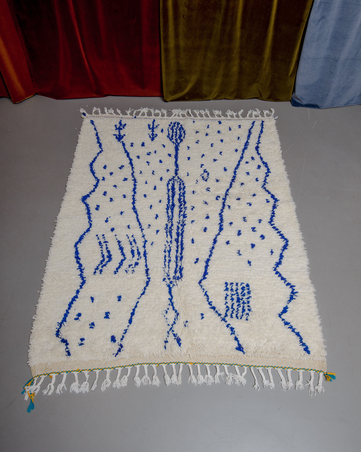 Azilal rug with abstract design in blue with yellow and turquoise details at one end  and white cotton tassels, laying on grey floor in front of velvet curtains. 