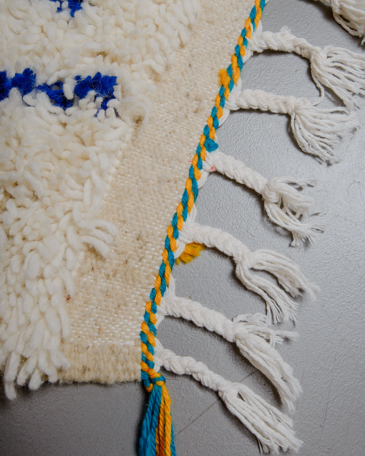 Corner of azilal rug with white cotton tassels and yellow and turquoise accents on grey floor. 