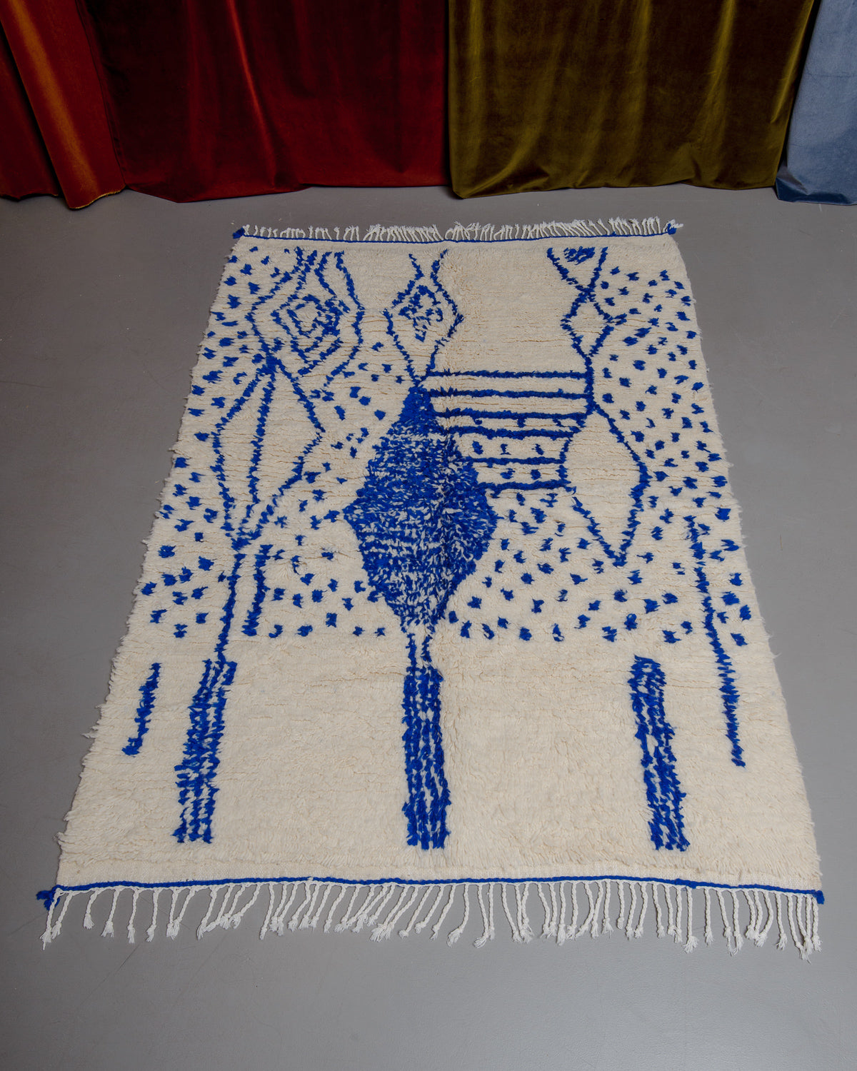 Azilal rug with abstract design in blue with  white cotton tassels, laying on grey floor in front of velvet curtains. 