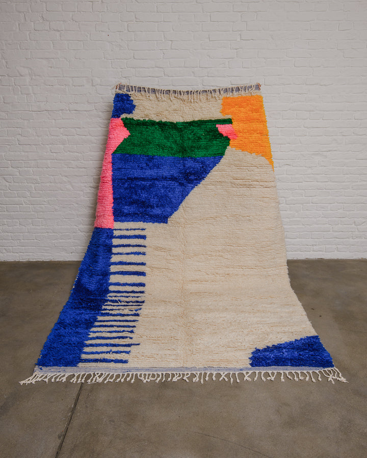 Cream azilal rug with bold forms in blue, green, pink and yellow being held up in front of a white brick wall. 