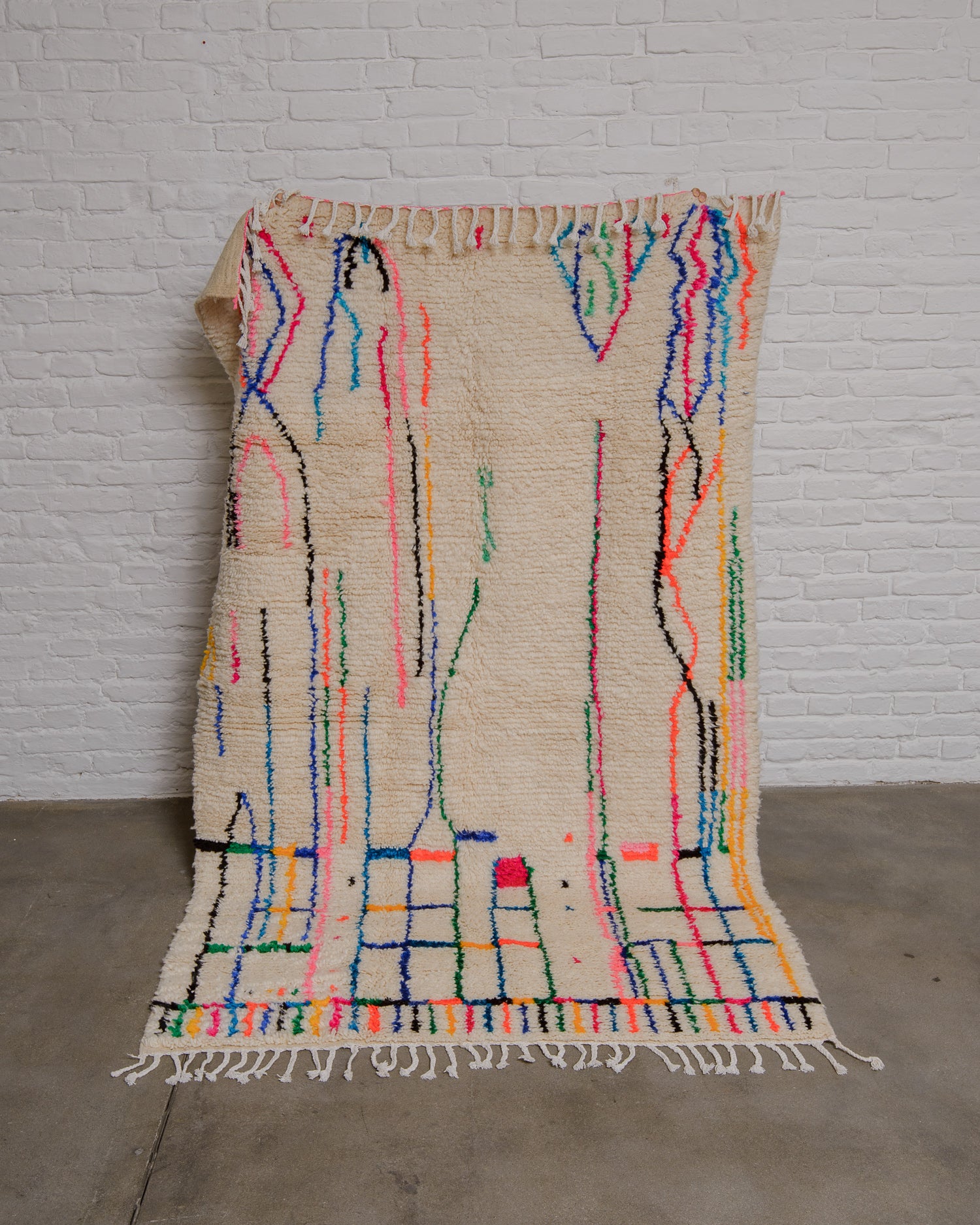 Cream azilal rug with colourful lines being held up in front of a white brick wall. 