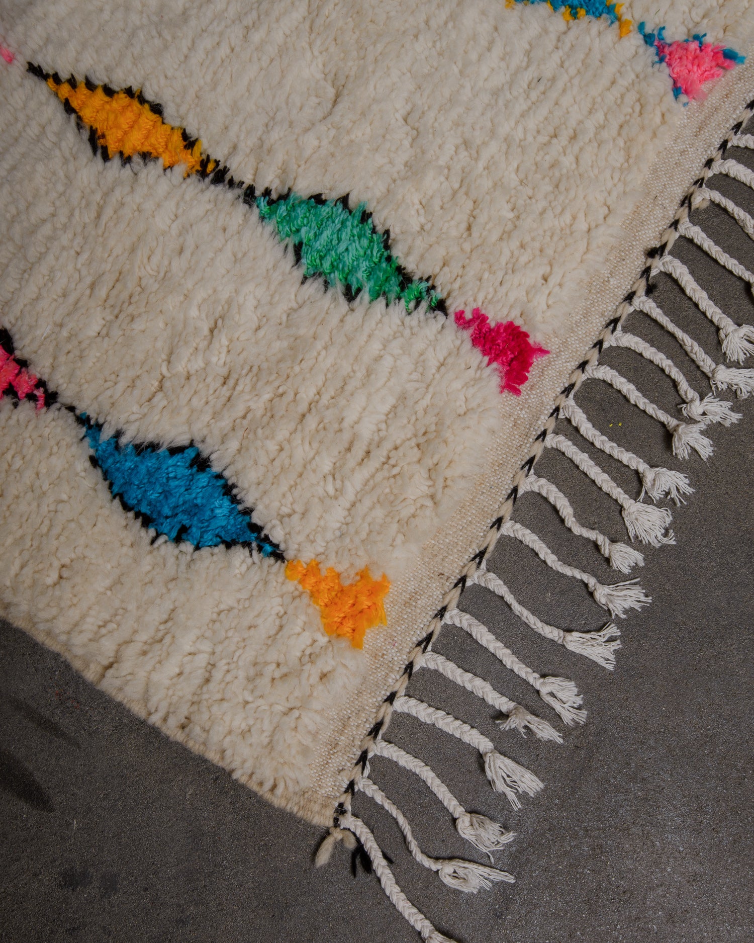 Corner of cream azilal rug with white cotton tassels on concrete floor. 