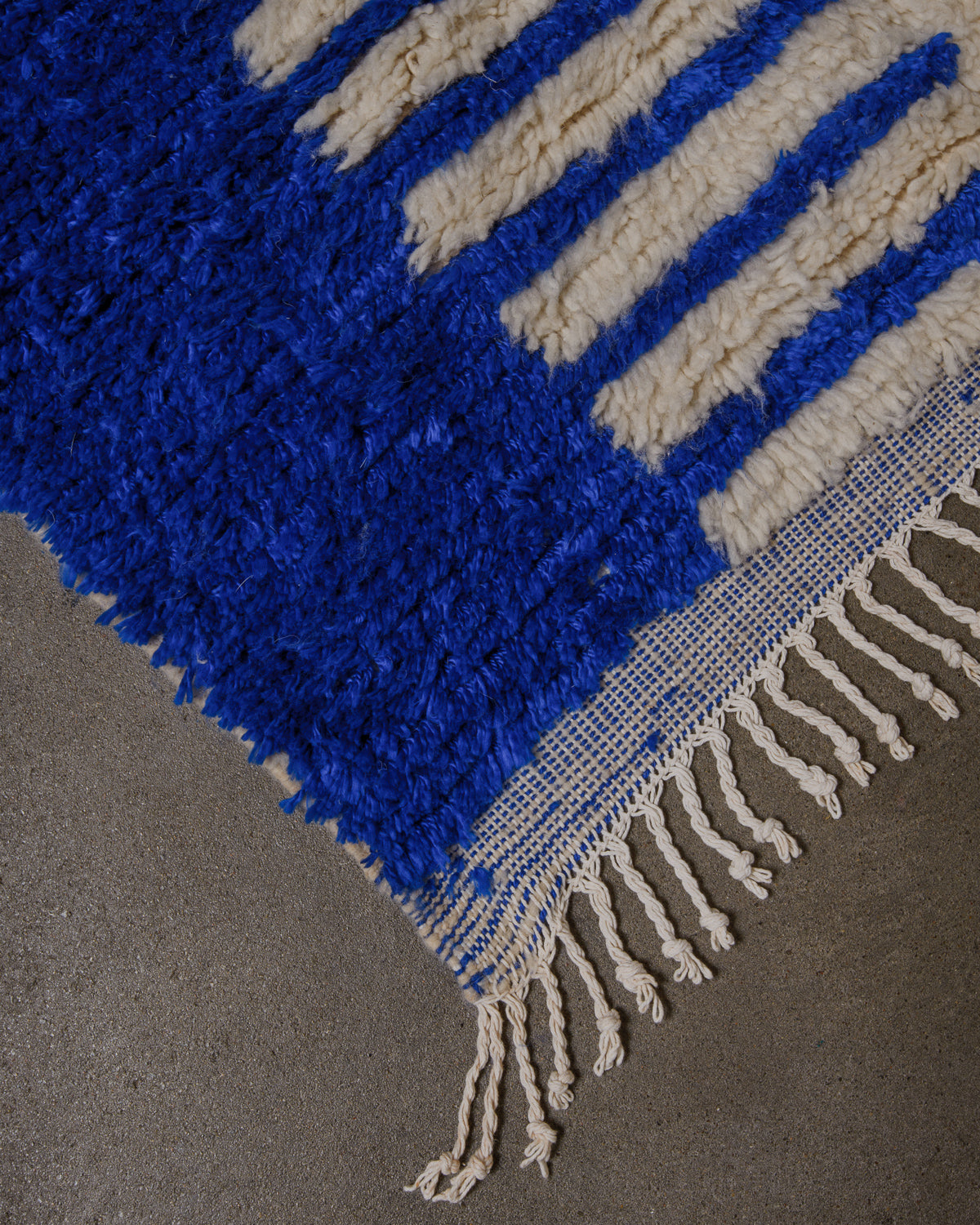 Corner of azilal rug with blue and cream, with white cotton tassels on concrete floor. 