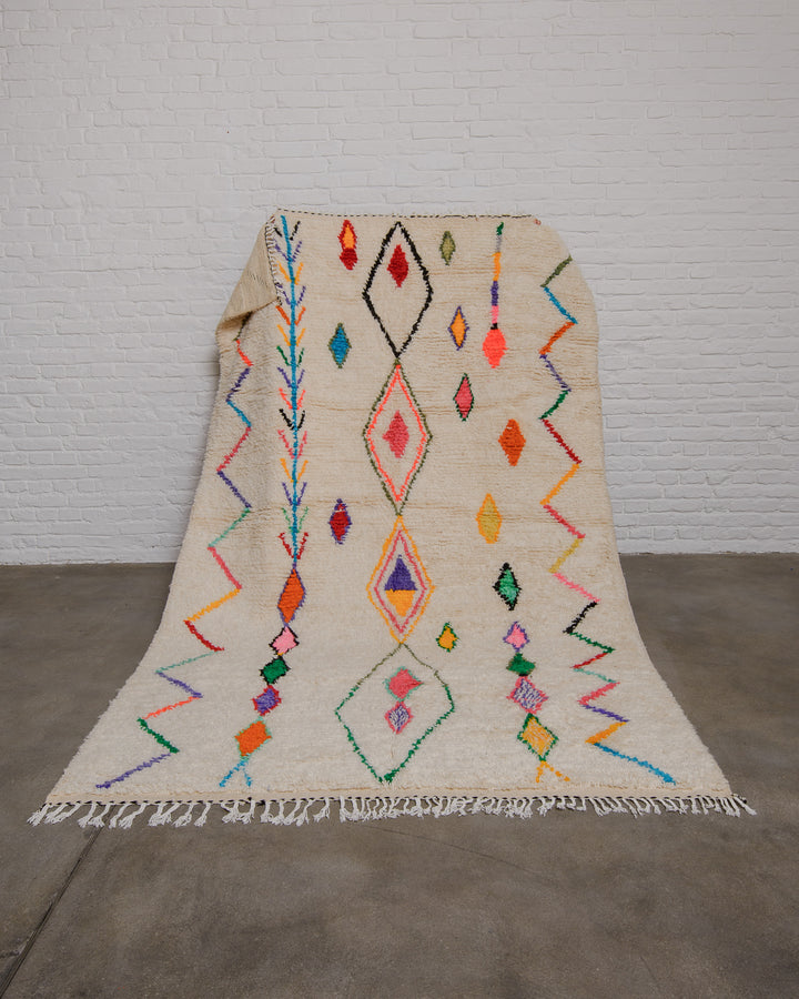 Cream azilal rug with colourful design elements being held up in front of white brick wall.