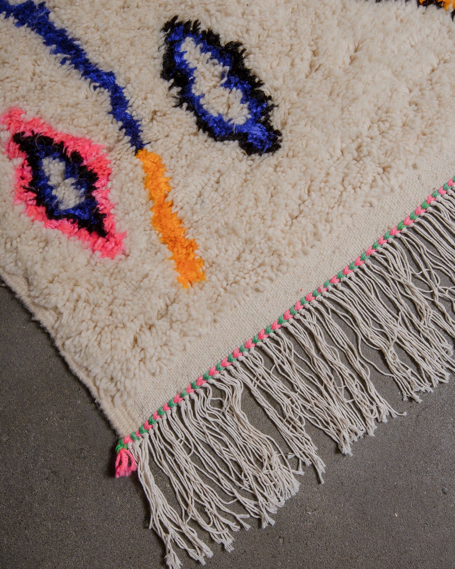 Corner of azilal rug made of cream wool with white cotton tassels and pink and turquoise stitching along the edge.