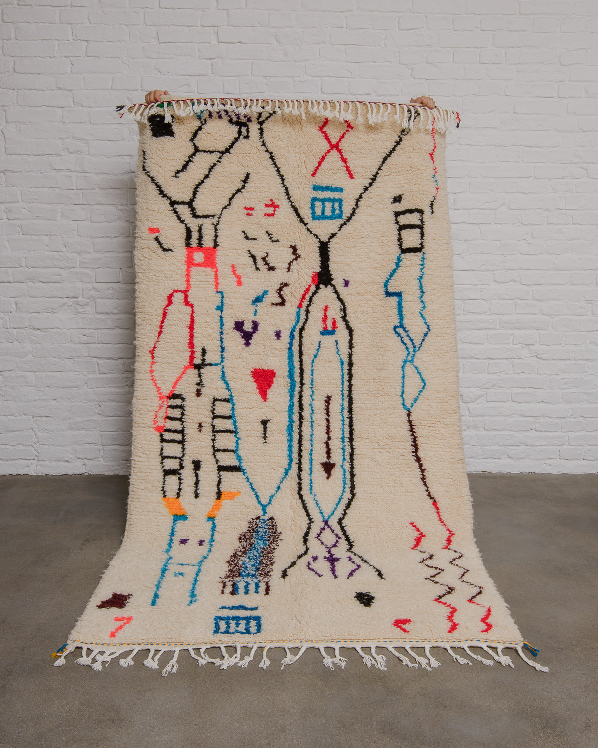 Cream Azilal rug with bold abstract design in red, black, blue, brown and purple being held up in front of a white brick wall. 