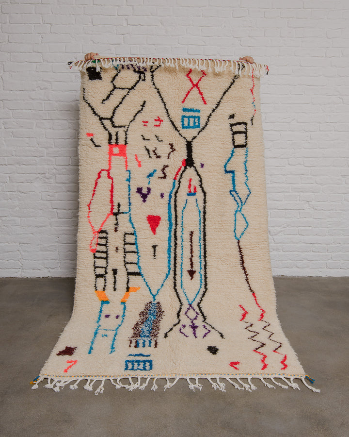 Cream Azilal rug with bold abstract design in red, black, blue, brown and purple being held up in front of a white brick wall. 