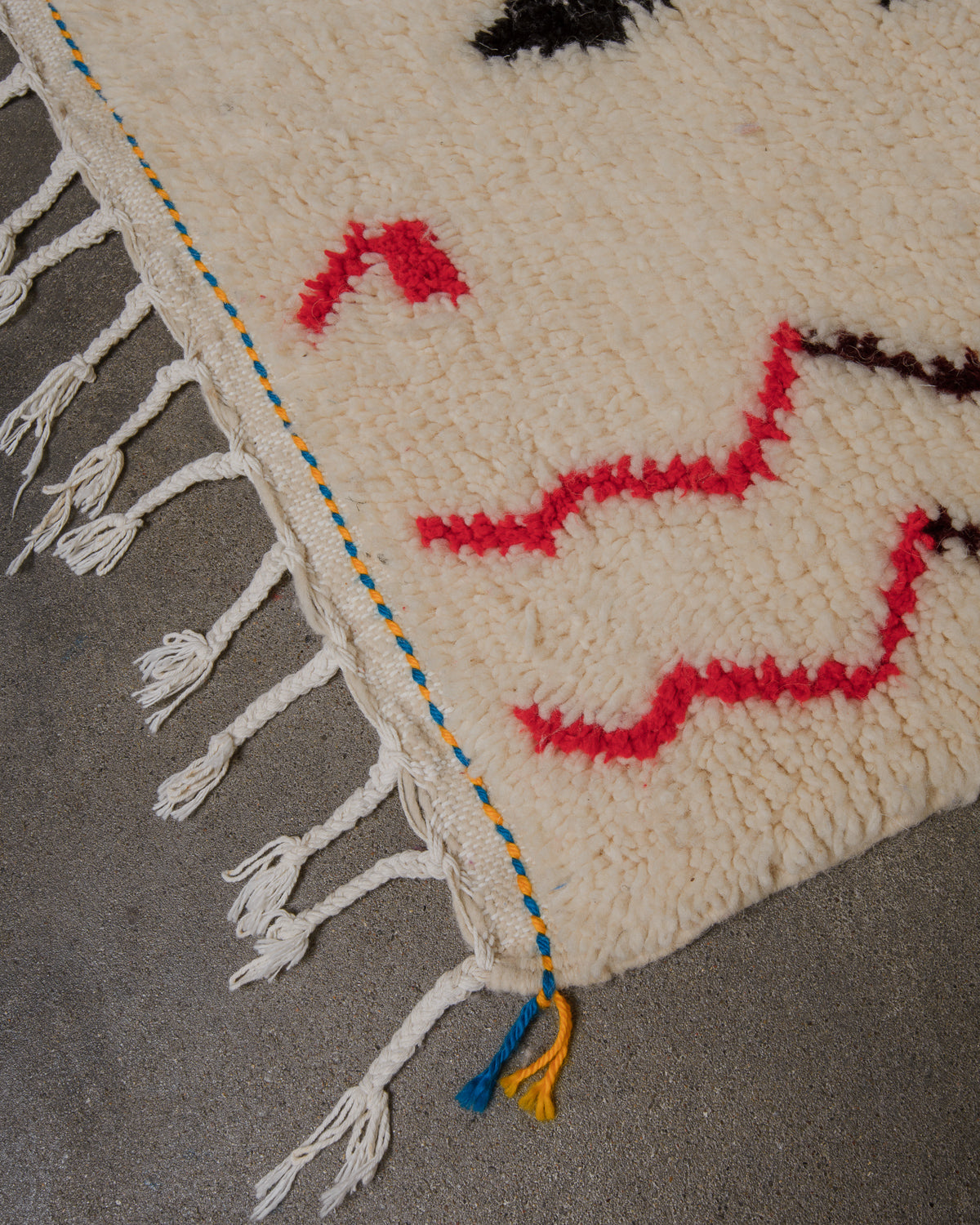 Corner of cream azilal rug with blue and yellow trim and white cotton tassels, on concrete floor. 