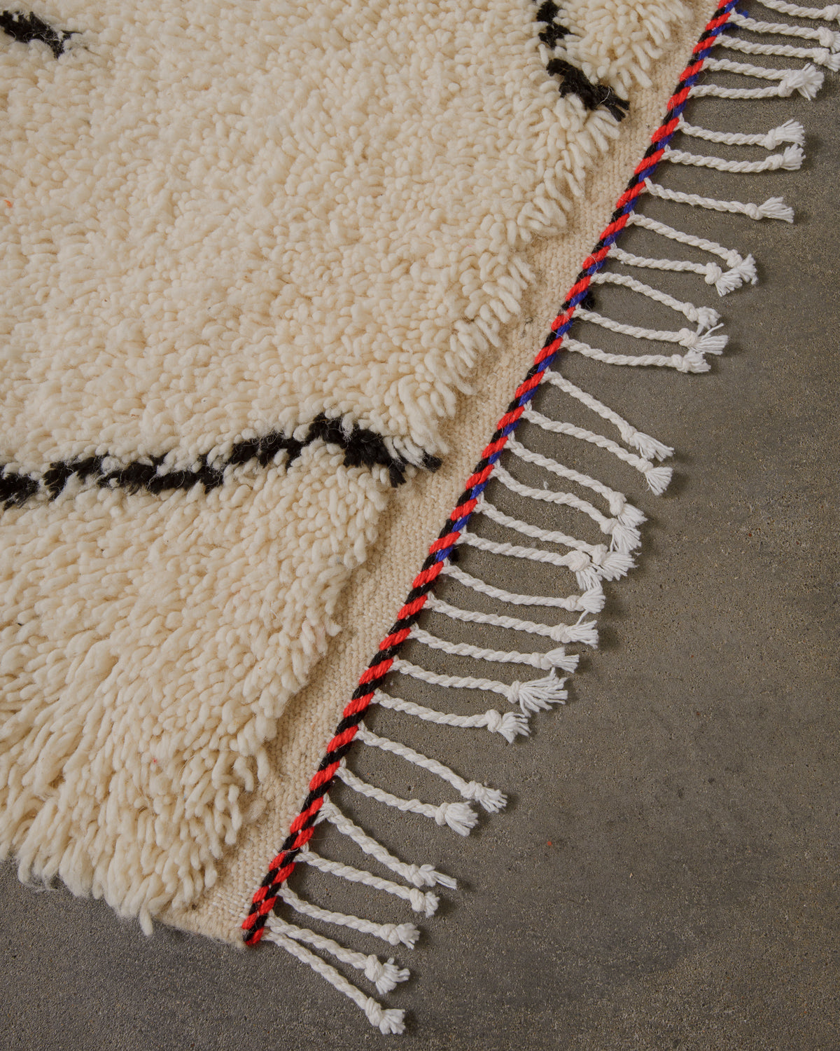 Corner of cream azilal rug with simple black design and black, blue and red trim across the end and white cotton tassels on concrete floor. 
