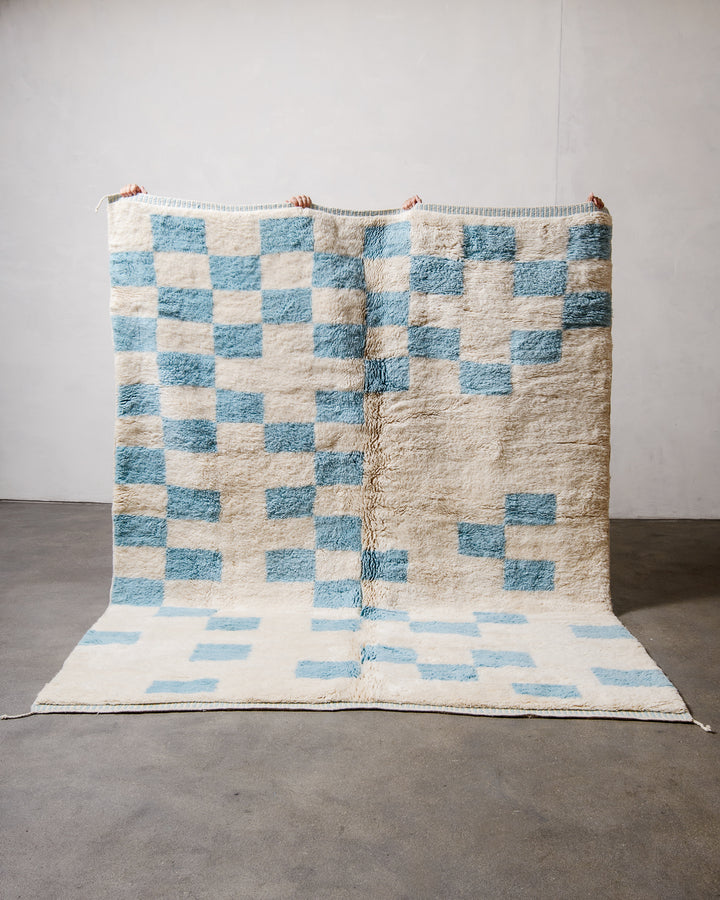 Berberlin Beni M'rirt Rug made of heavy wool pile in cream and sky blue being held up in front of a white wall and concrete floor. 