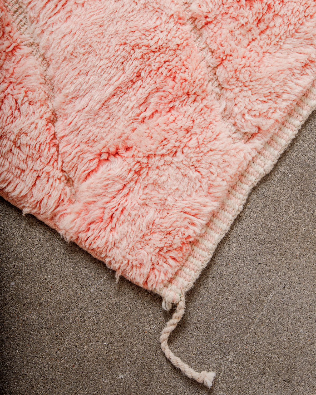 Corner of rug made of thick wool pile in coral pink and finished with tightly woven end with alternating lines of pink and cream, and a single wool tassel on the corner. 