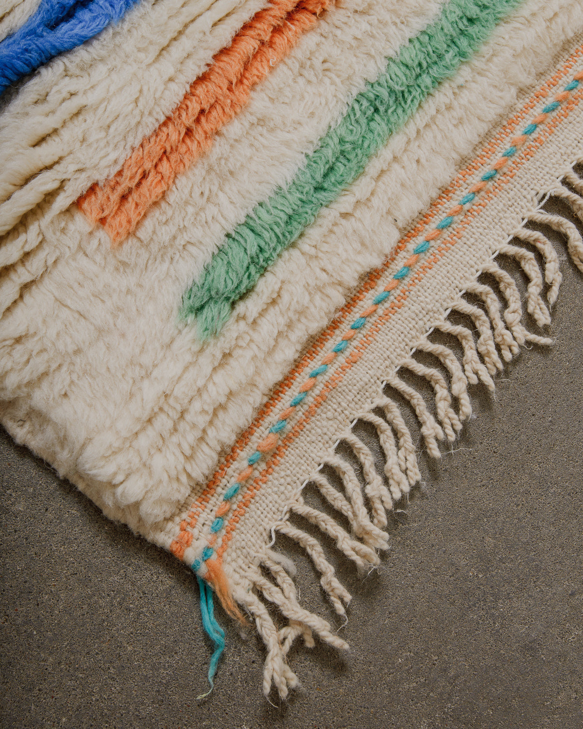 Corner of wool rug with woven design across the end and wool tassels on a concrete floor. 