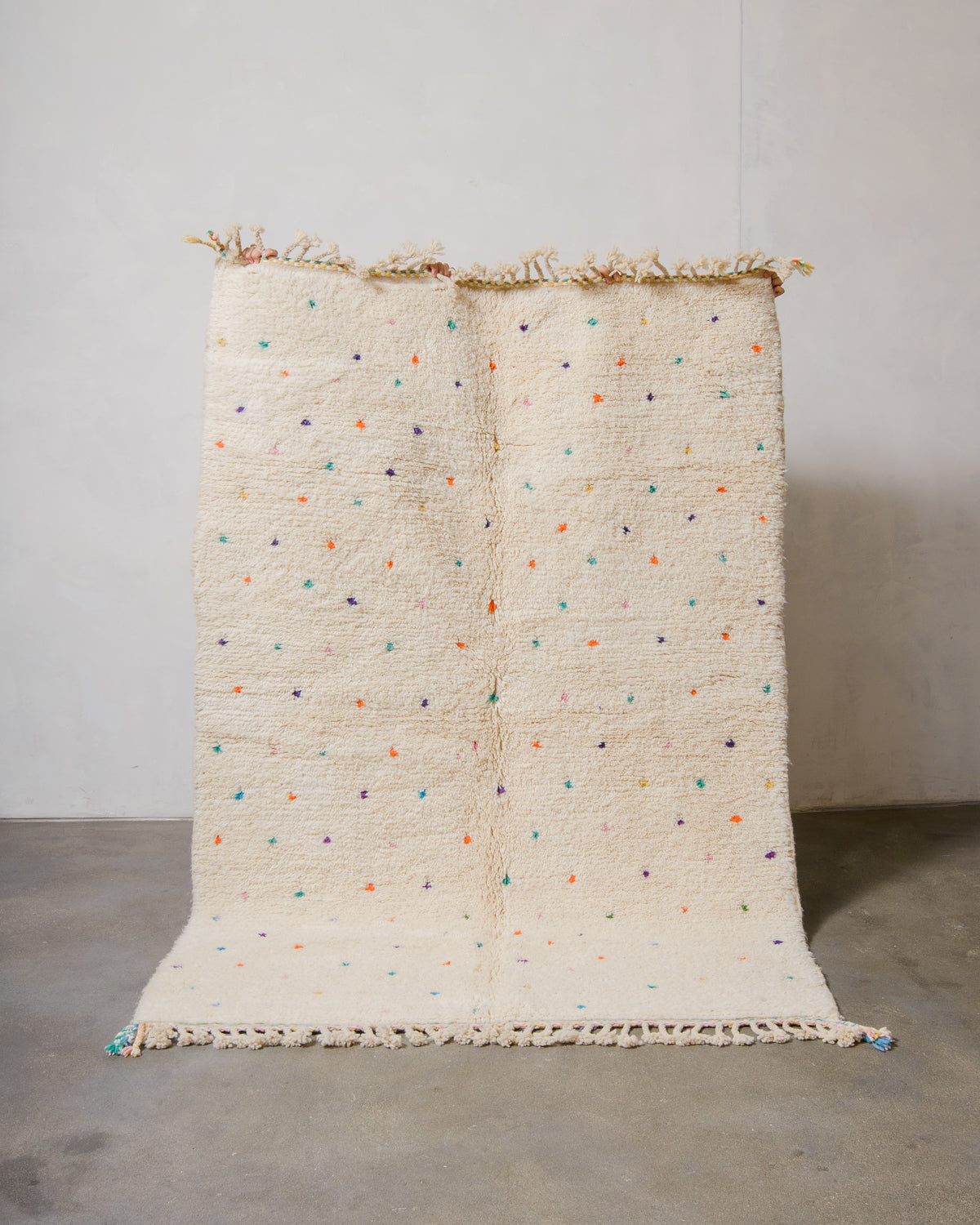Berberlin Beniourain rug in cream wool with coloured dots being held up in front of a white wall. 