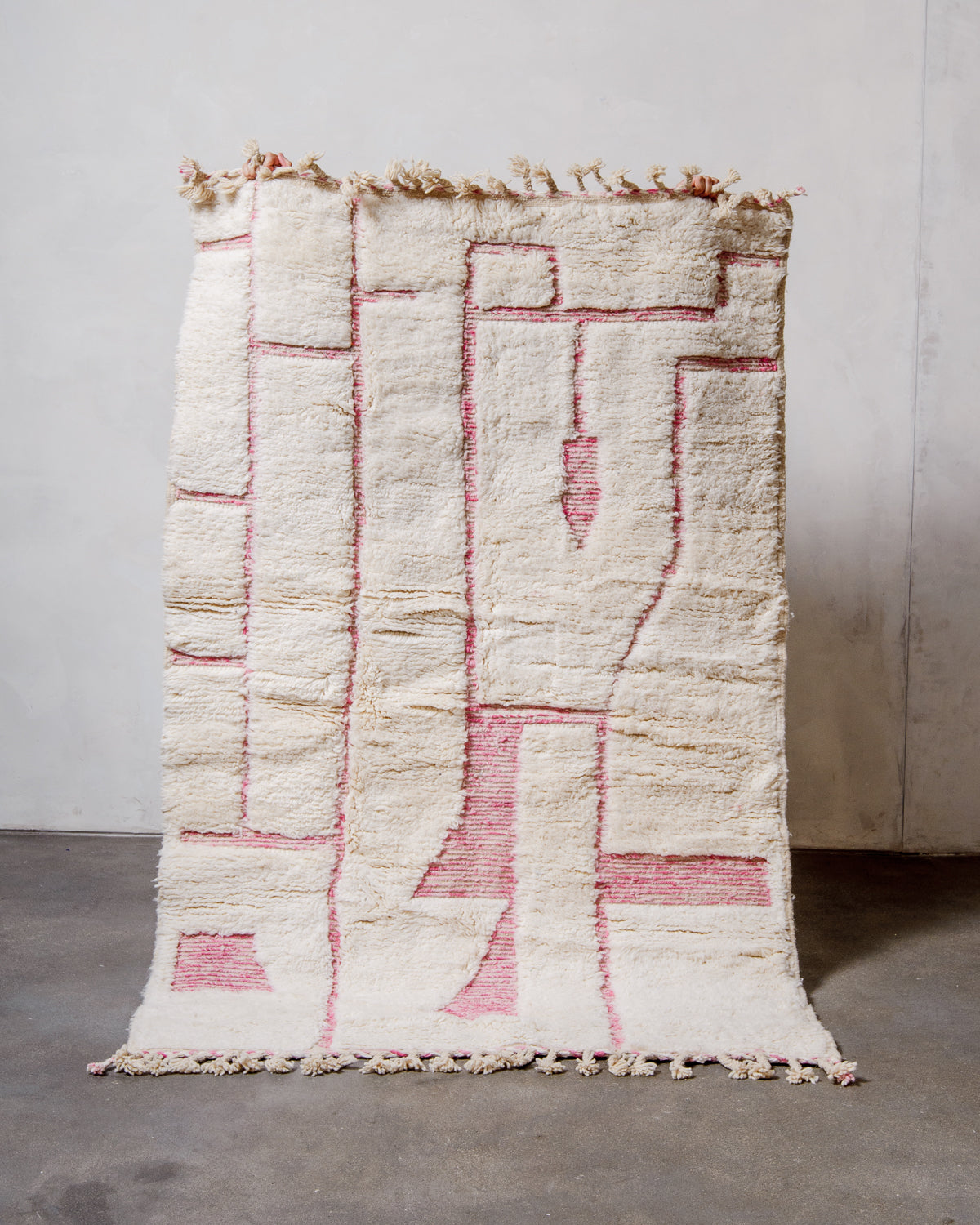 Berberlin Beniourain rug in cream and pink wool being held up against a white wall and a concrete floor. 