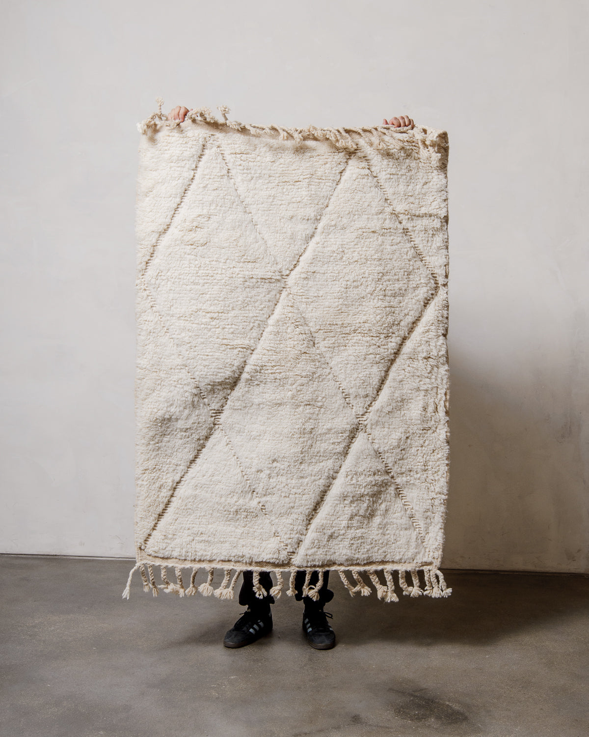 Small Berberlin Beniourain rug in cream wool with diamond design, being held up in front of a white wall. 