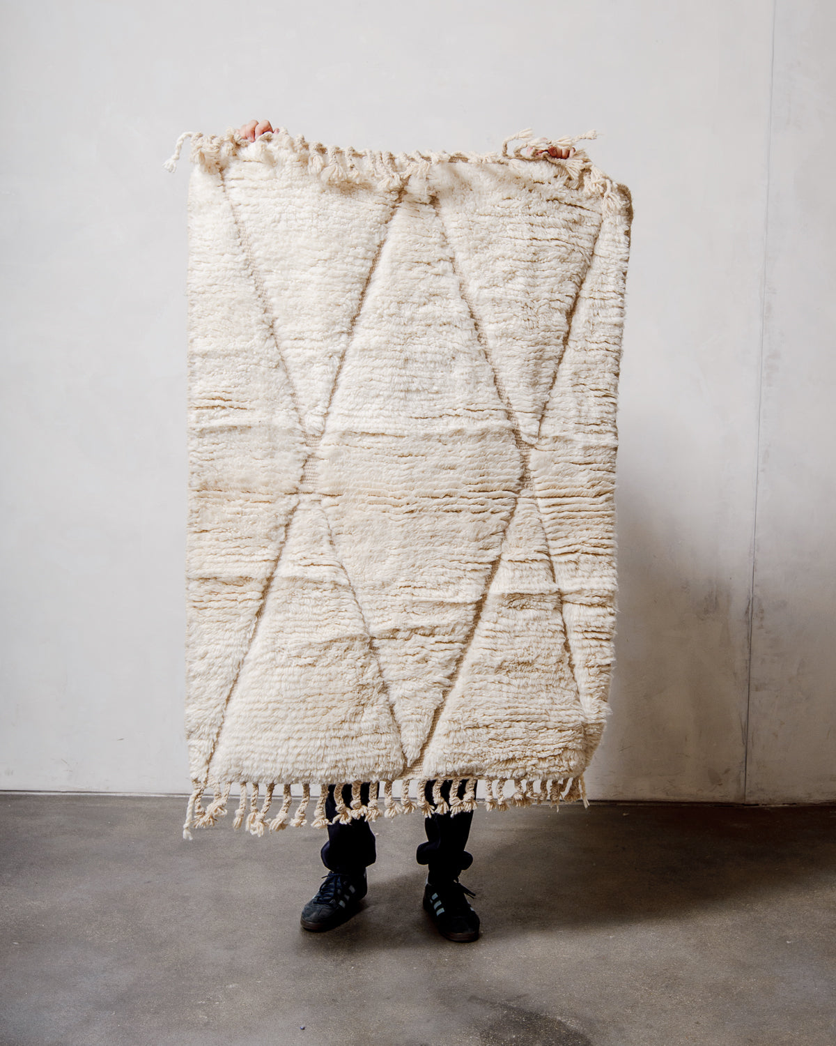 Small Berberlin Beniourain rug in cream wool with diamond design, being held up in front of a white wall. 