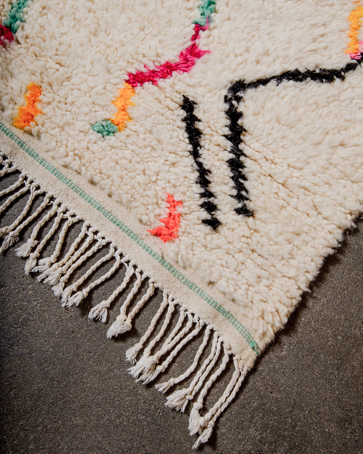 Corner of cream Azilal rug with a tightly woven edge and white cotton tassels across the end. 