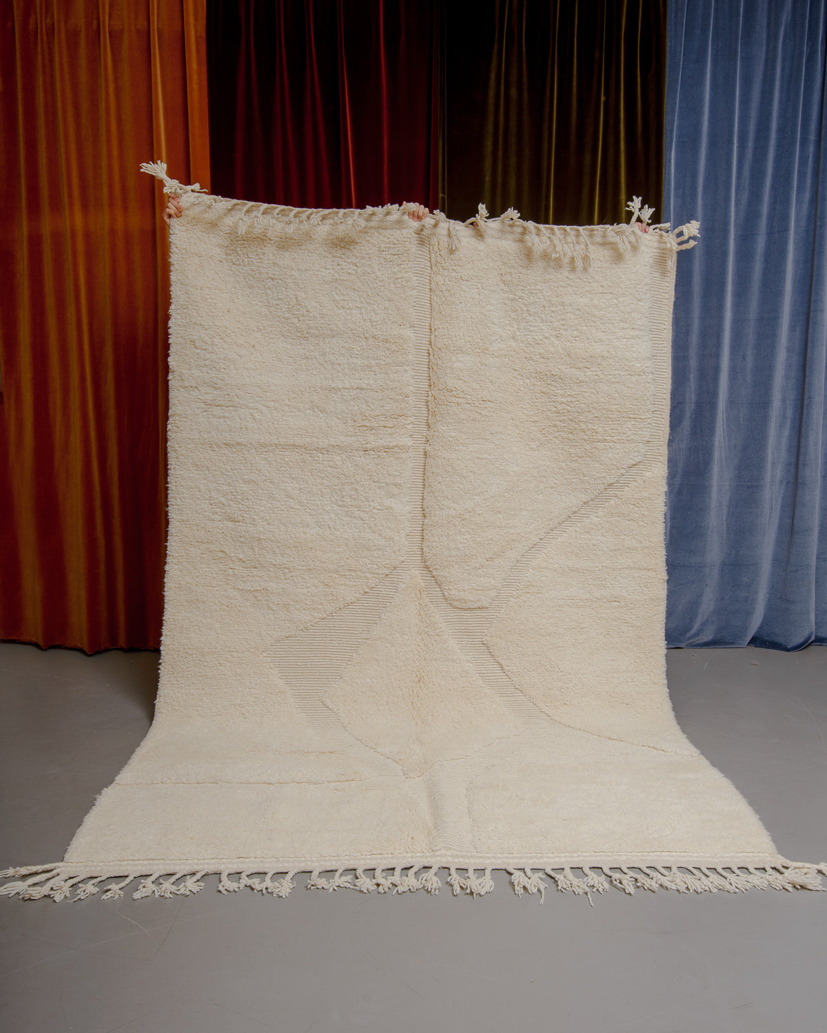 Berberlin Beniourain rug in cream wool with abstract pattern being held up in front of velvet curtains.