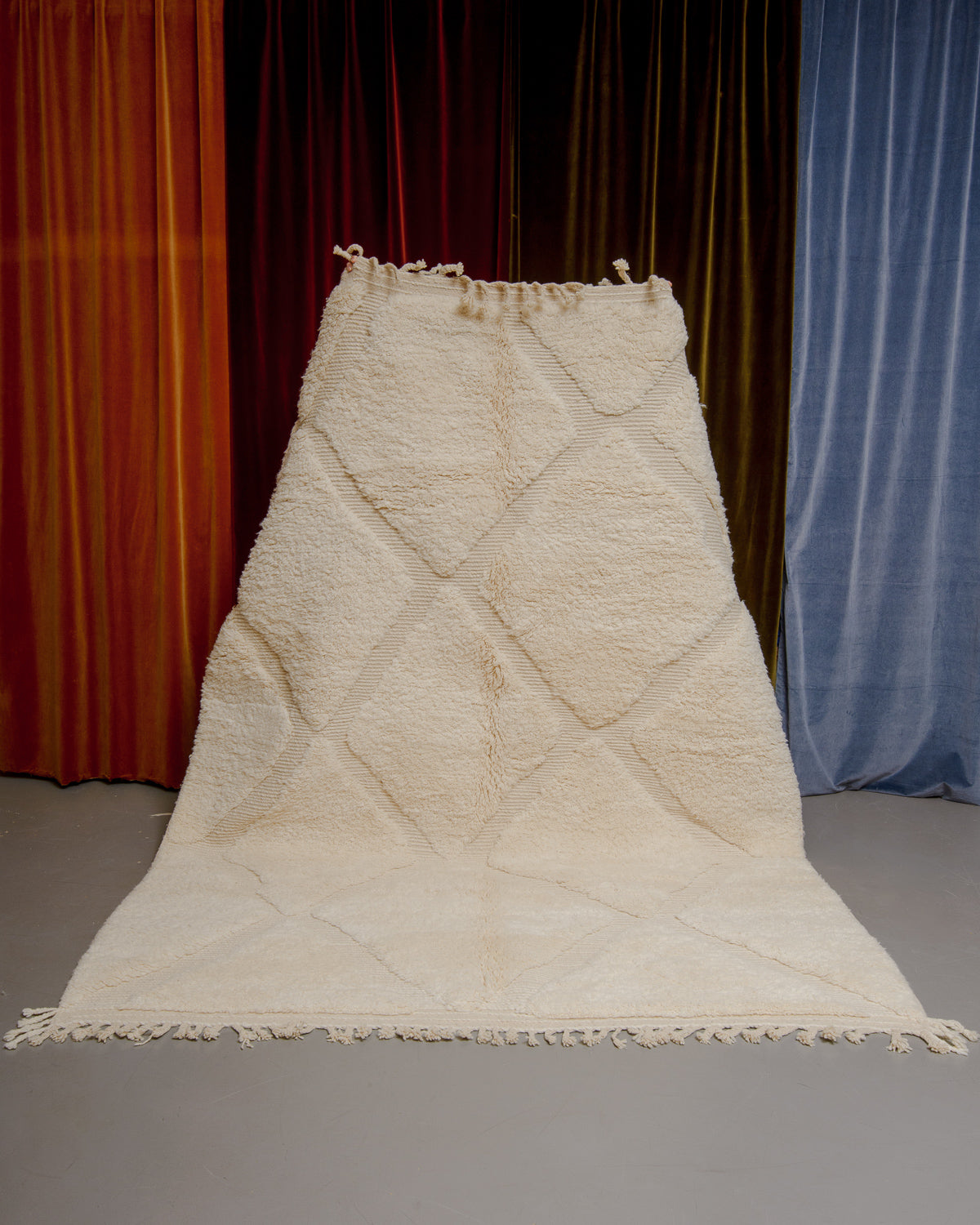 Berberlin Beniourain rug in cream wool with diamond pattern being held up in front of velvet curtains.