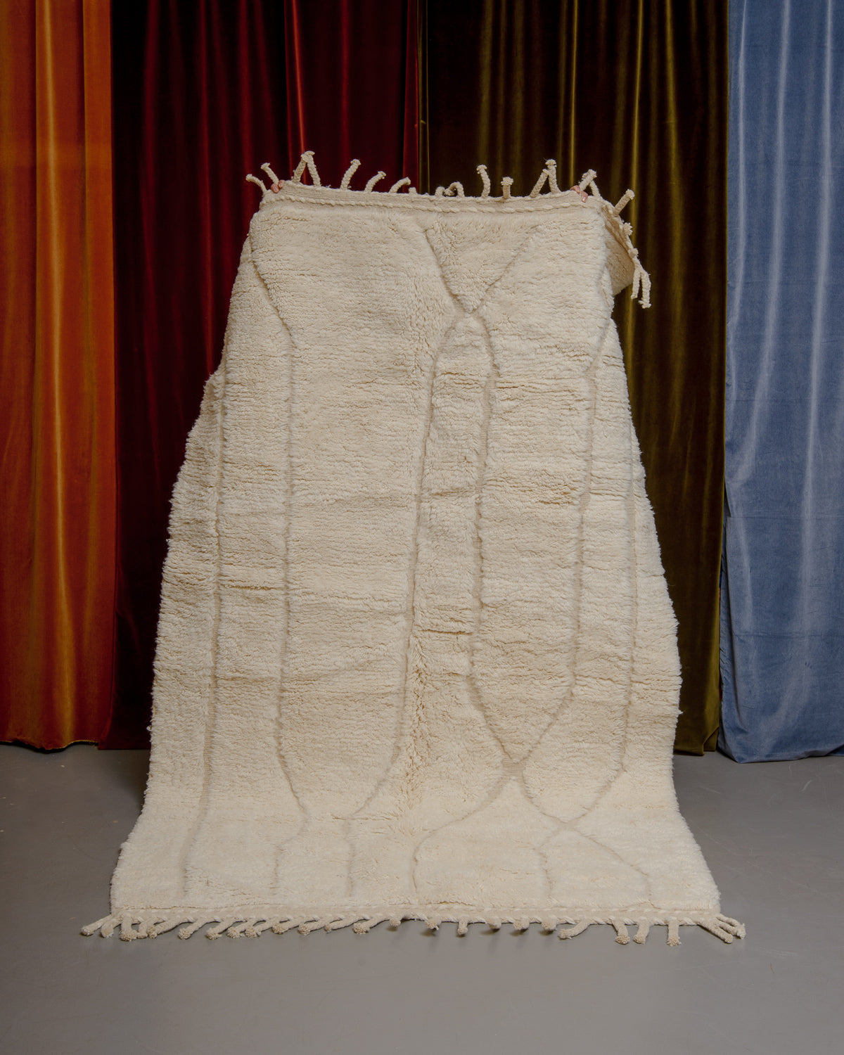 Berberlin Beniourain rug in cream wool with abstract pattern being held up in front of velvet curtains.