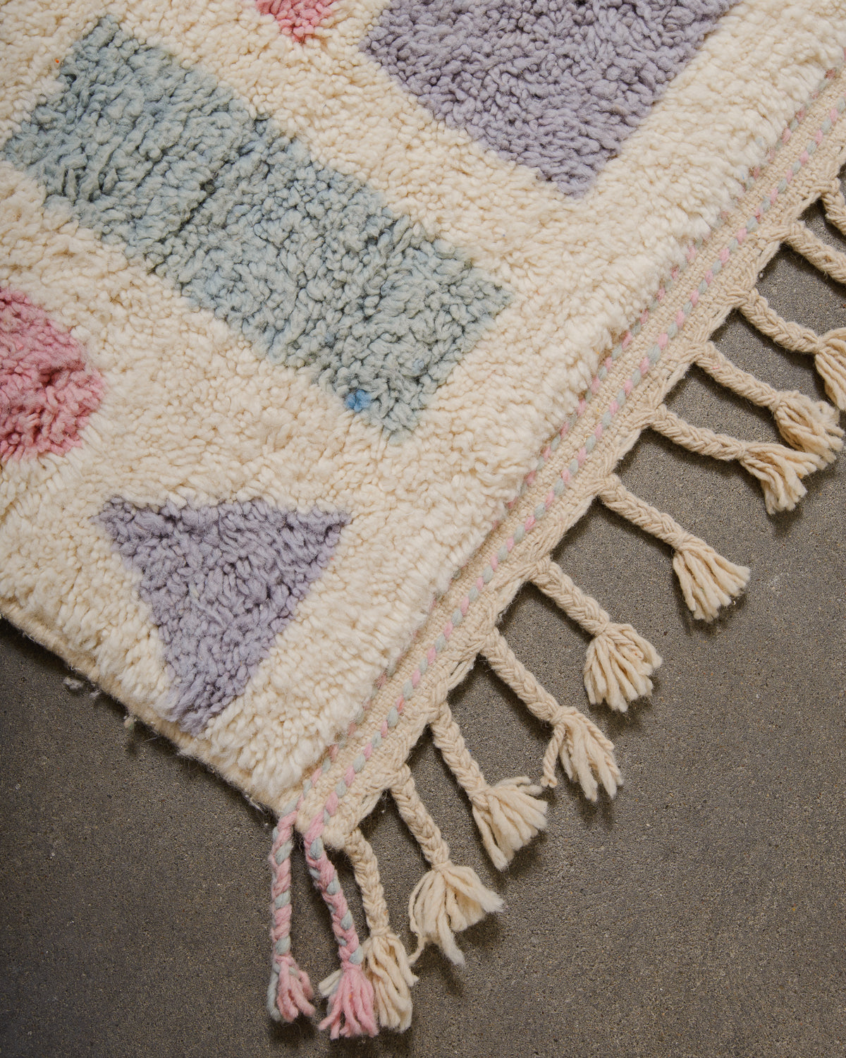 Corner of berber rug that is finished with tightly woven ends with pastel pink and blue details and natural wool tassels across the ends.