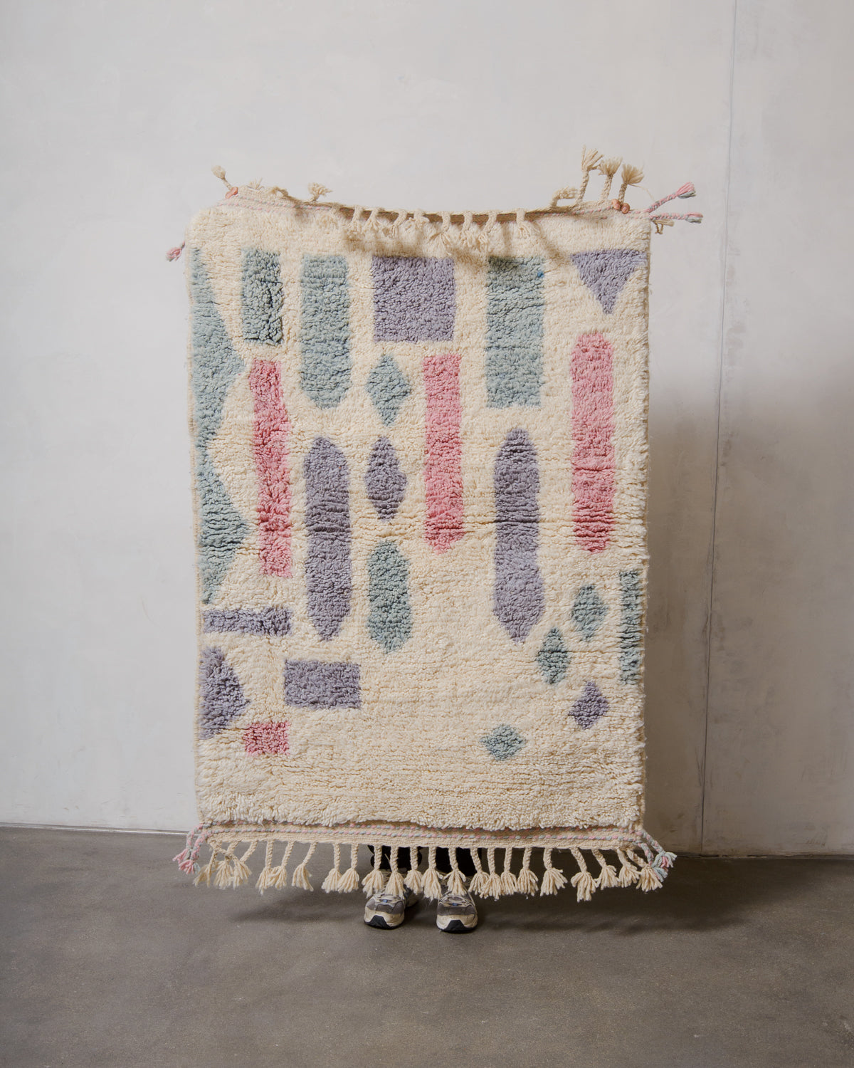 Small Berberlin Beniourain rug made of  cream and pastel coloured wool, being held up in front of a white wall. 