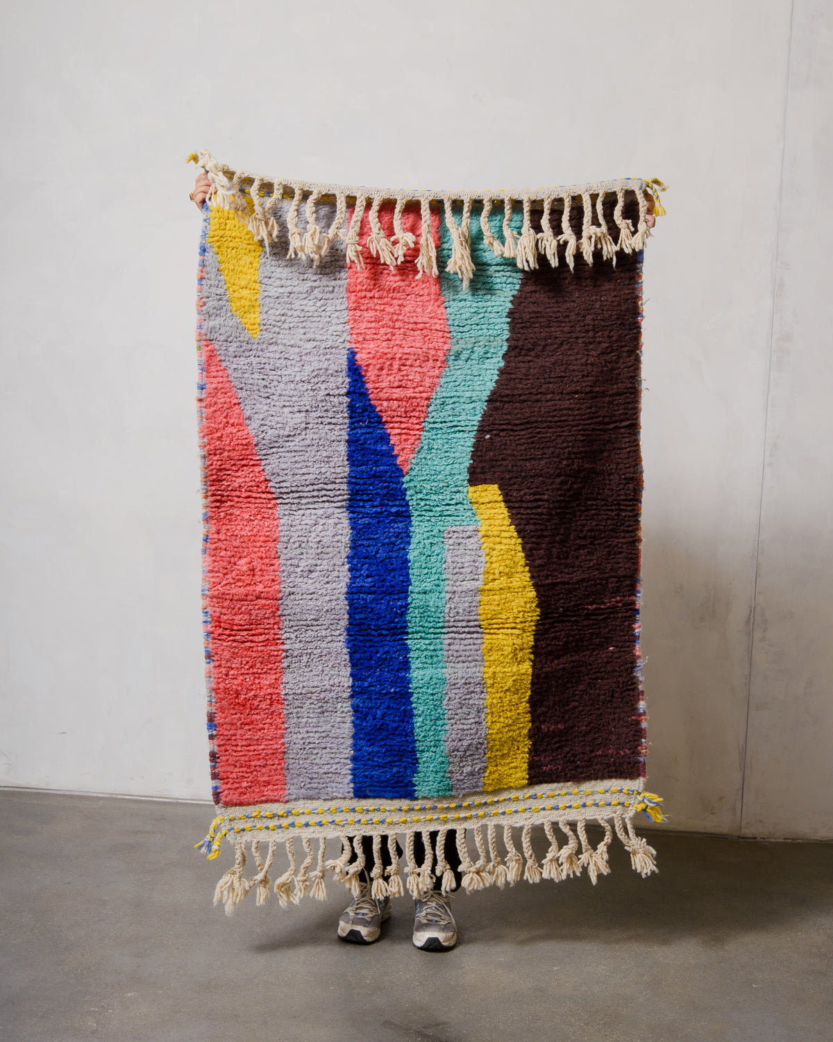 Small Berberlin Beniourain rug made of  colourful wool, being held up in front of a white wall. 