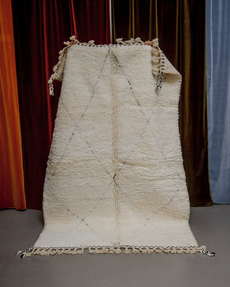 Berberlin Beniourain rug in cream wool with black diamond pattern being held up in front of velvet curtains.