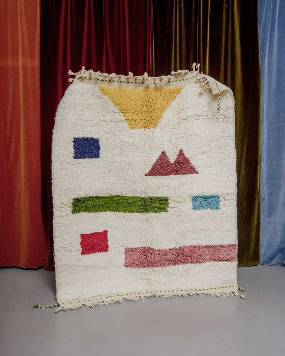 Berberlin beniouran rug in cream with colourful forms being held in front of velvet curtains.