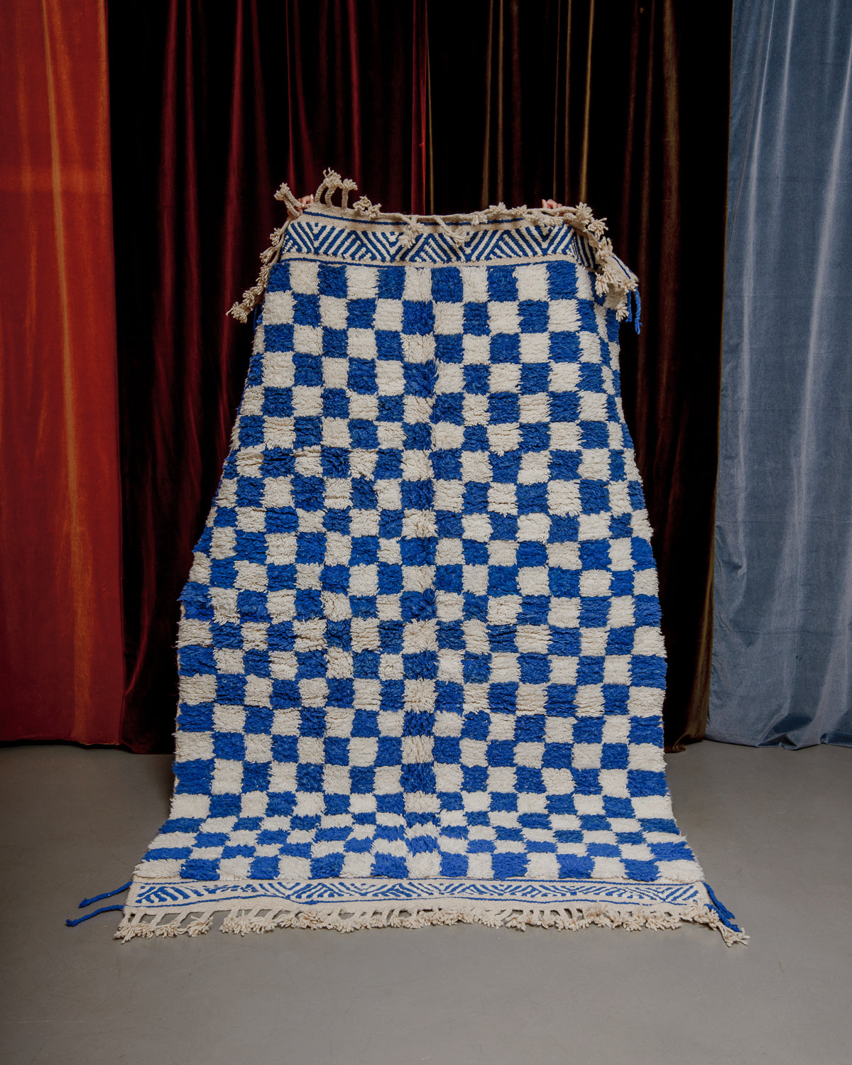Berberlin beniourain rug in blue and white checkers with patterned border, being held in front of velvet curtains.