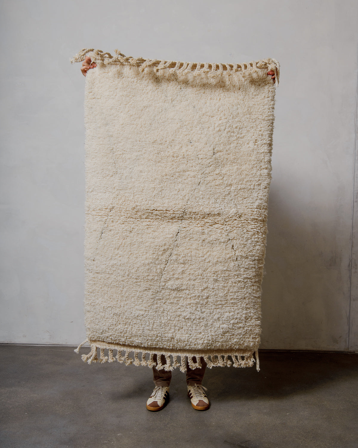 Textured cream wool rug with tassels held by a person against a plain wall.