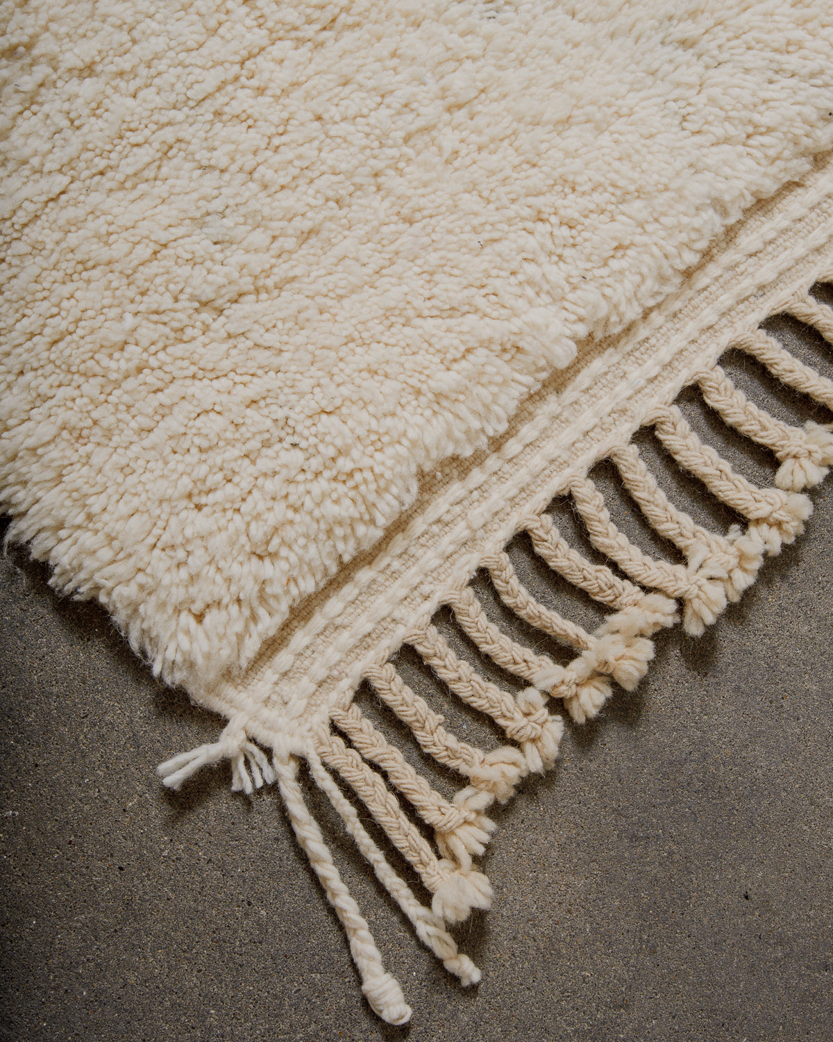 Corner of cream Berberlin beniourain rug with natural wool tassels on concrete floor.
