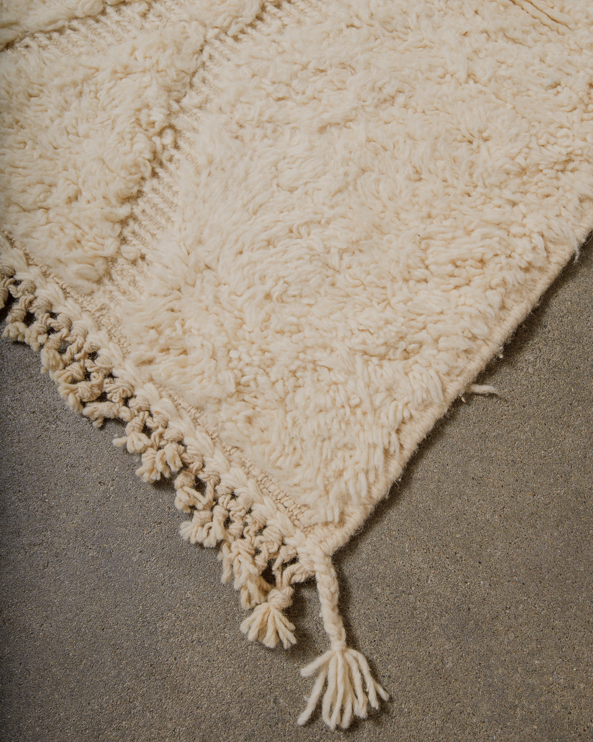 Corner of Berberlin beniourain rug in cream wool with natural wool tassels on a concrete floor.