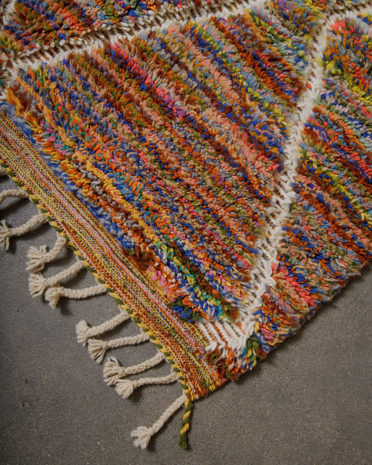 Multicoloured textured rug with tassels on a grey surface.