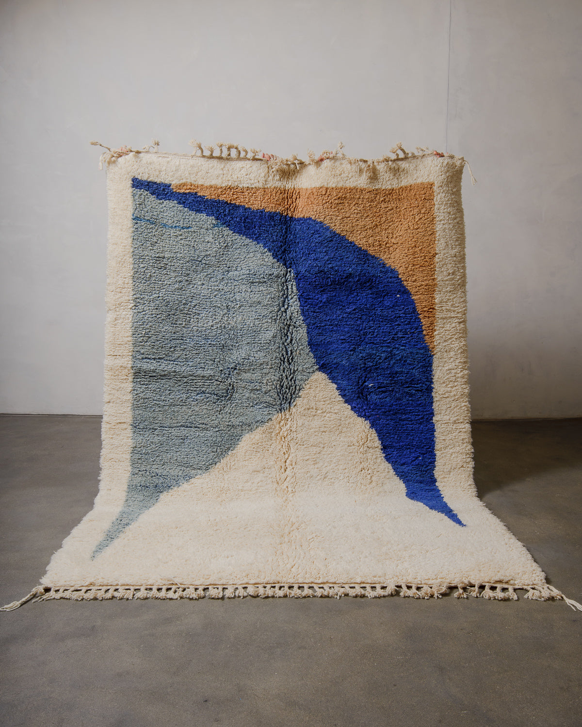 Beniourain rug with shades of blue, cream and camel, held up in front of a plain wall.