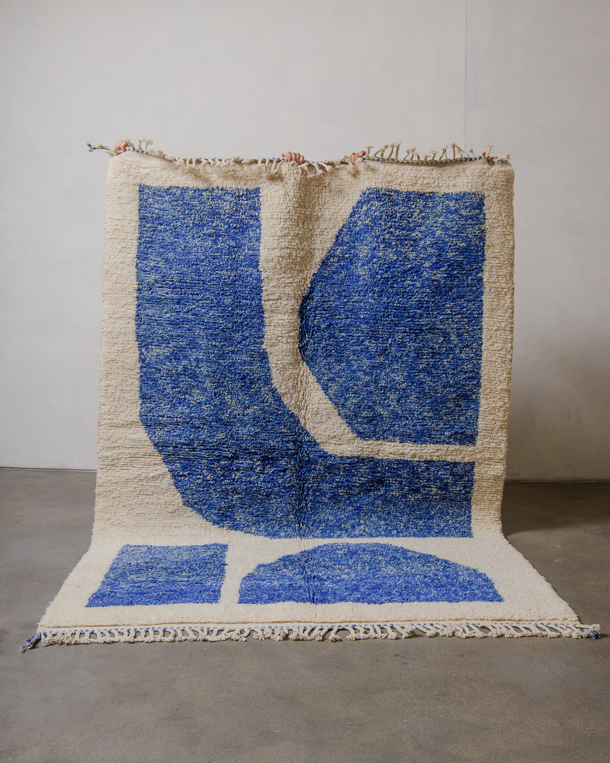 Berberlin Rug with bold blue design held up in front of a plain wall. 