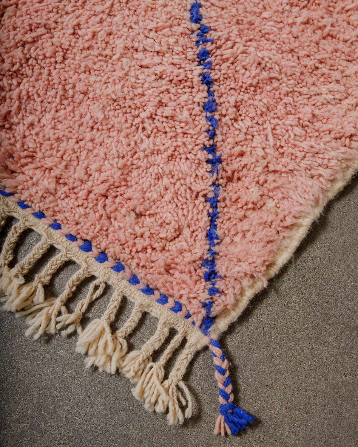 Corner of wool beniourain rug in pink and blue with natural wool tassels on concrete floor. 