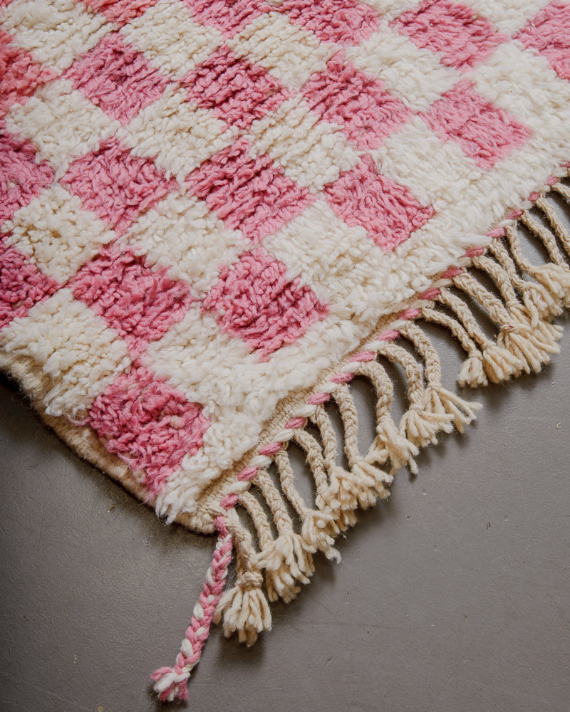 Corner of pink checkered rug with natural wool tassels and pink and cream edge. 
