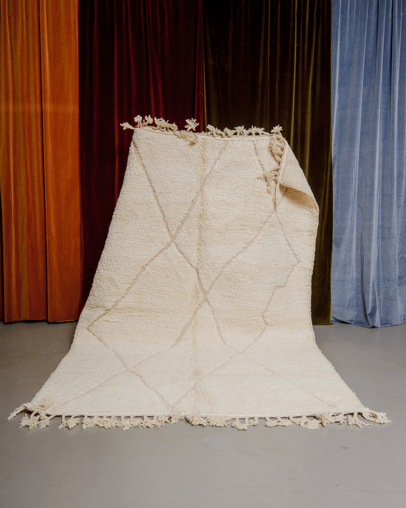 Berberlin Beniourain rug in cream wool being held up in front of velvet curtains.