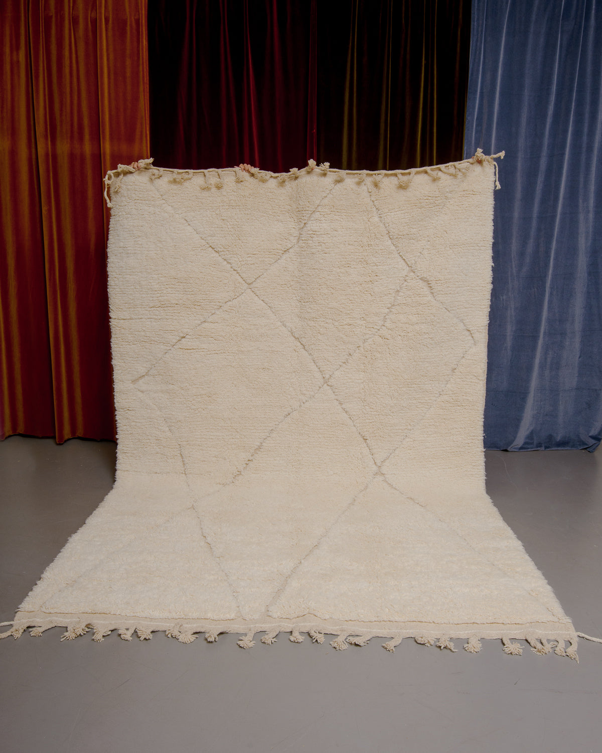 Berberlin Beniourain rug in cream wool with diamond pattern being held up in front of velvet curtains.