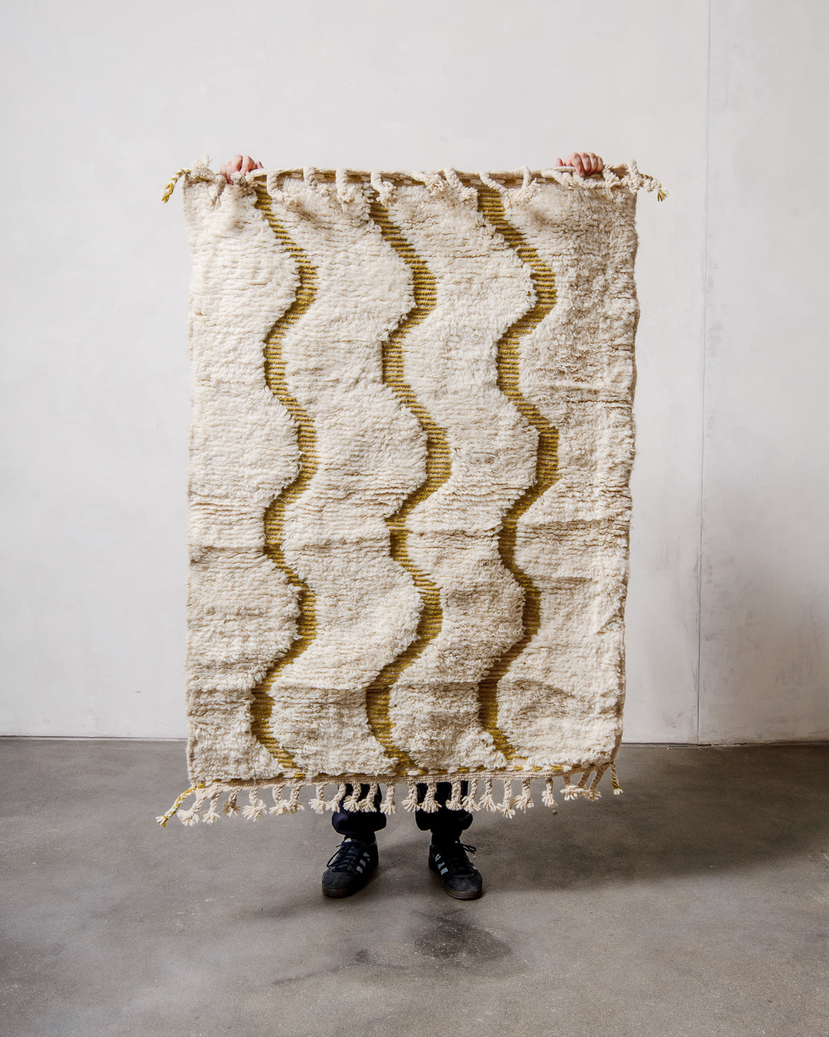 Small wool Berberlin Beniourain rug in cream featuring woven olive green waves, being held up in front of a white wall. 