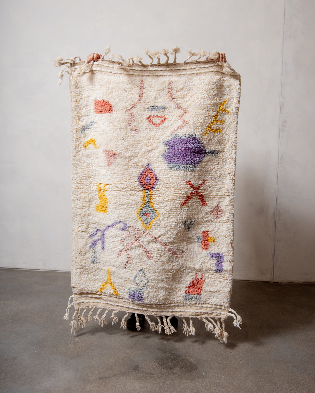 Small Berberlin Beniourain rug made of cream wool with pastel coloured abstract design, being held up in front of a white wall. 