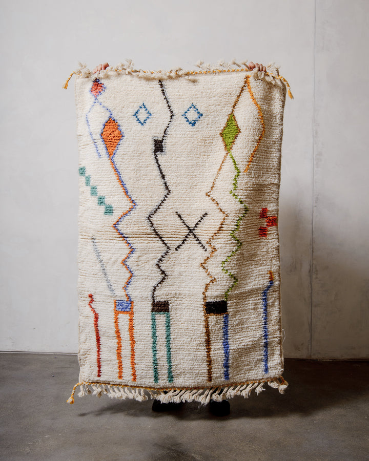 Small Berberlin Beniourain rug made of cream wool with colourful, abstract design, being held up in front of a white wall. 