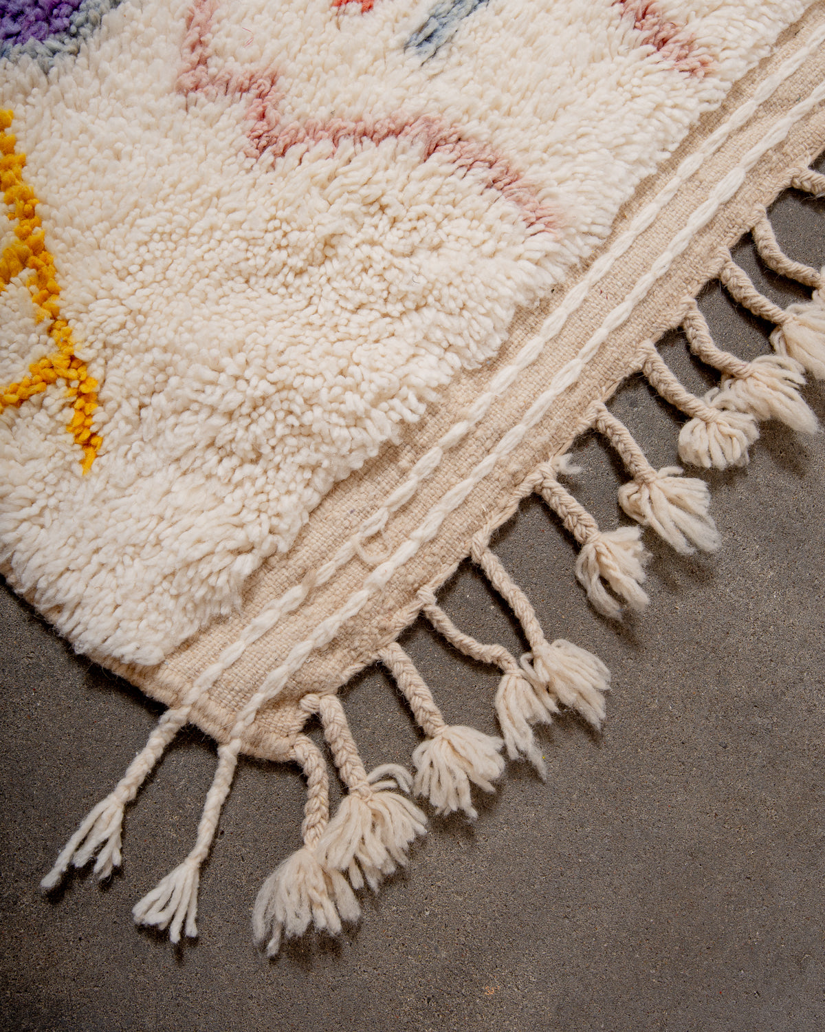 Corner of cream berber rug that is finished with tightly woven ends and natural wool tassels.