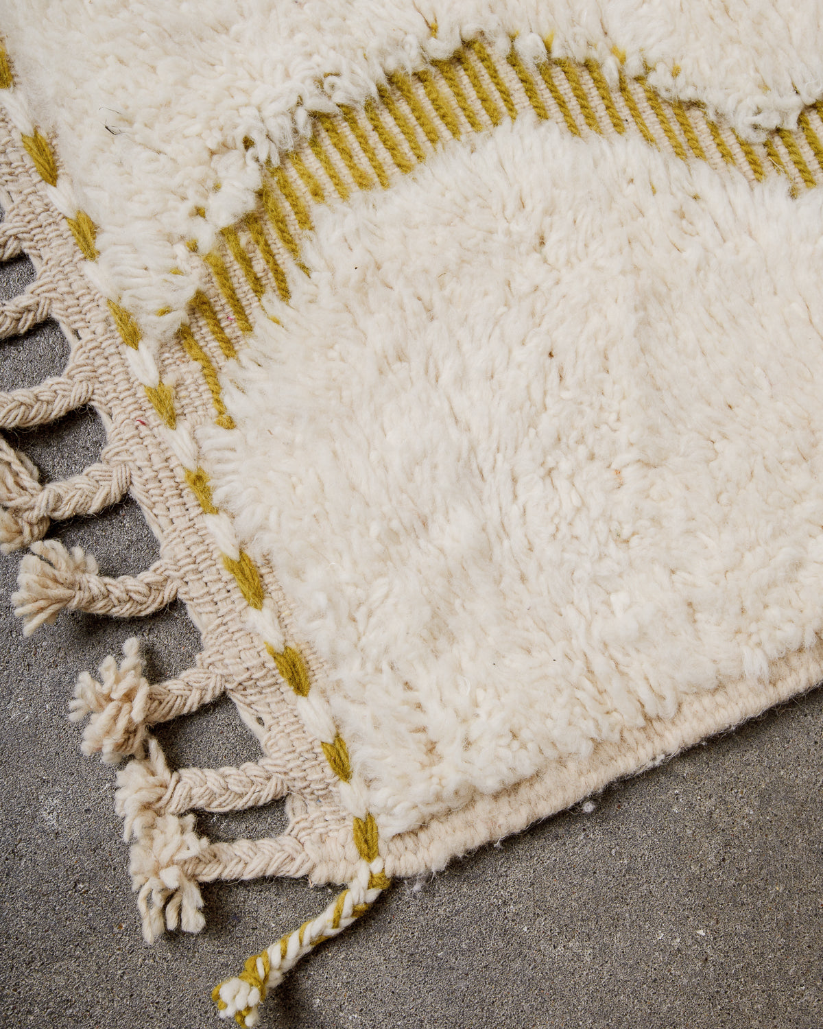 Corner of cream Beniourain wool rug with a tightly woven edge and natural wool tassels laying on concrete floor. 