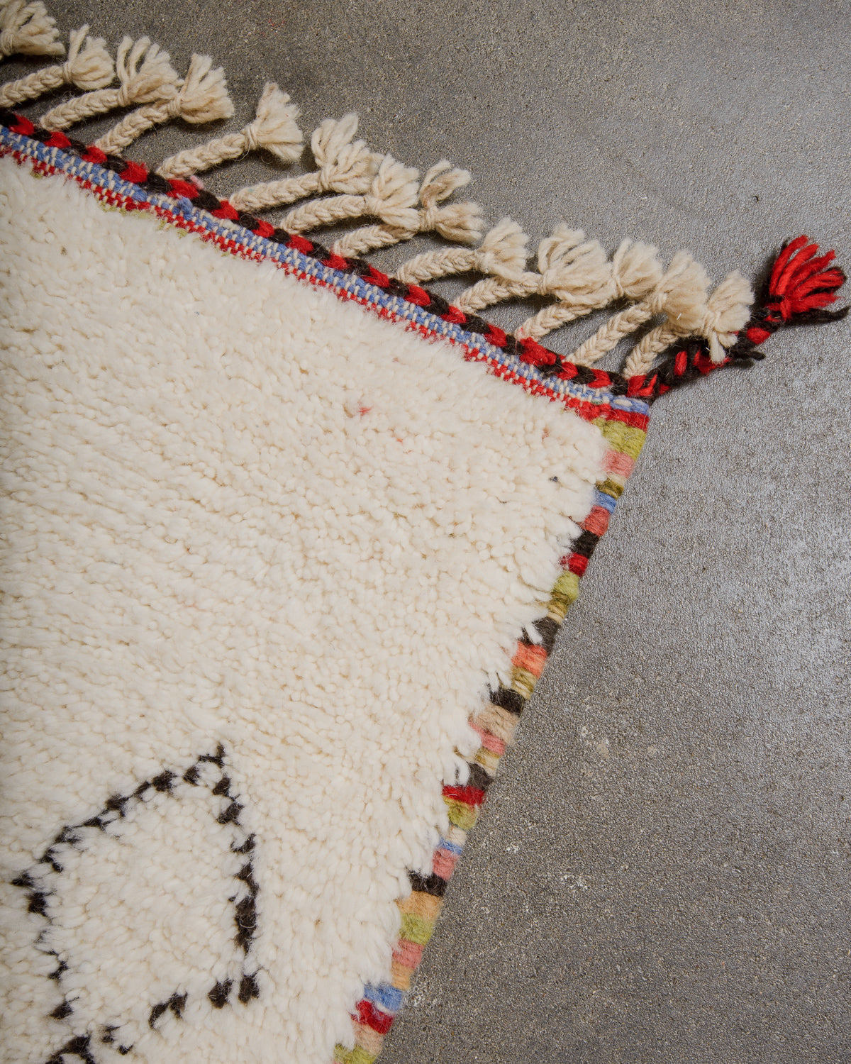 Corner of rug with red and black trim and natural wool tassels and multicoloured border laying on concrete floor. 