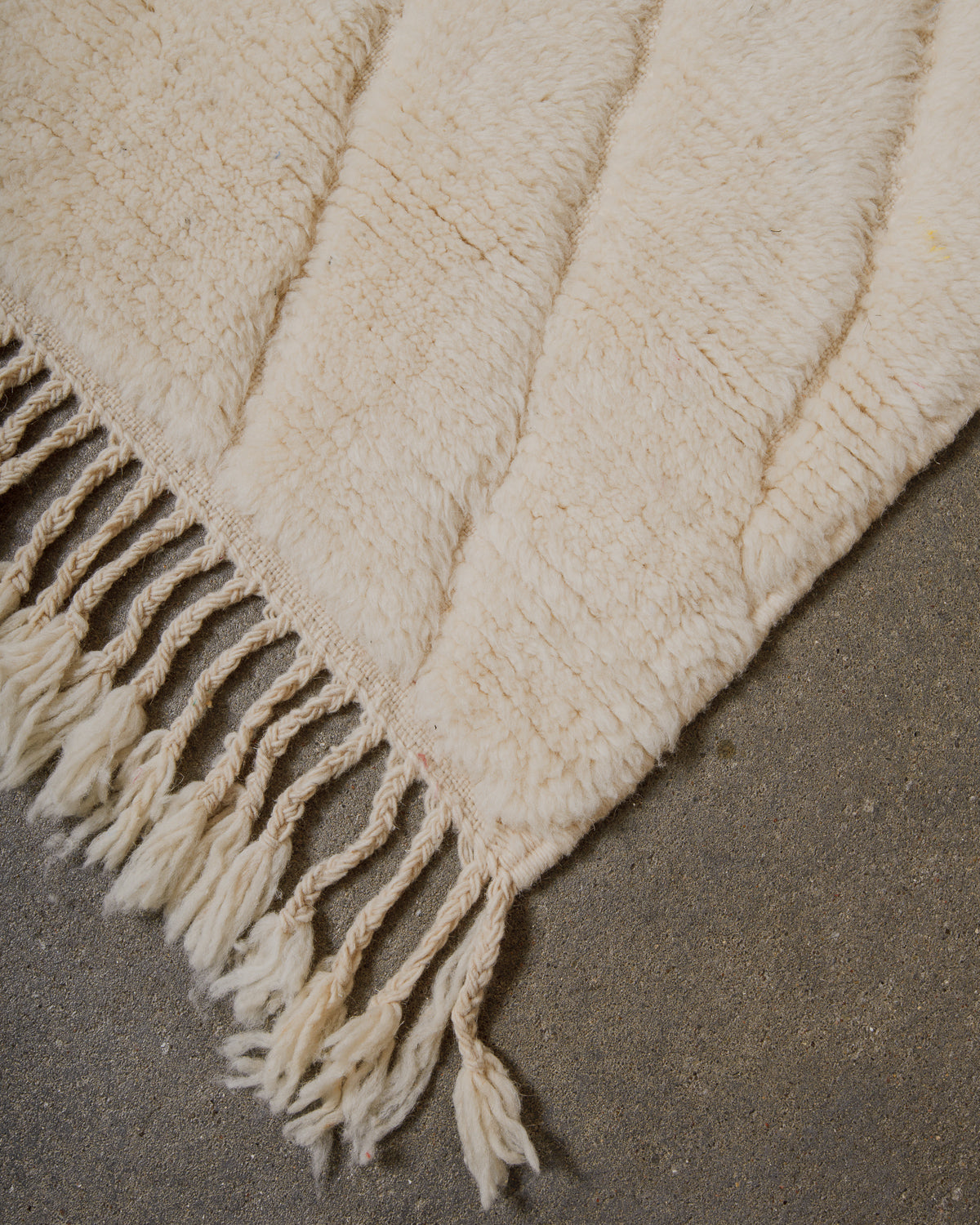 Corner of cream wool rug with tassels on a concrete surface.